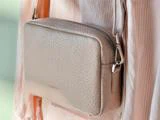 Nærbillede af en crossbody-taske i lys beige tekstureret læder med lynlås, båret over et lyserødt, plisseret outfit.