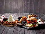 En saftig burger med salat, tomat og ost ligger ved siden af løgringe, stegte snacks og en vaffel med softice på et rustikt træbord.