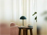 Et lille bord med en sort lampe, en kop kaffe og en bog står ved siden af en lyserød sofa foran hvide gardiner. En grøn plante er delvist synlig til højre.