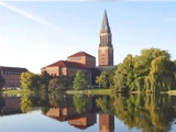 En stor murstensbygning med et højt klokketårn spejler sig i en rolig sø, omgivet af træer under en klar himmel.