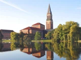En stor murstensbygning med et højt klokketårn står ved en rolig sø, hvor man kan se dens spejlbillede og de omkringliggende træer i vandet.