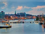 En livlig byhavn ved solnedgang med både, der ligger til kaj, moderne og historiske bygninger, en kirke med et grønt spir og byens skyline oplyst af lys.