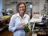 En kvinde med krøllet hår og briller står smilende i et køkken med en krukke og friske krydderurter i hånden. På komfuret ved siden af hende ses tilberedt mad på en pande.