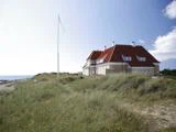 Et stort hus med rødt tag ligger blandt højt græs nær kysten under en klar blå himmel med en tom flagstang foran.