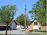En gårdsplads med bygninger, grønne træer, to flagstænger med flag, en stor sten, et 30 km/t-skilt og flere parkerede biler under en klar blå himmel.