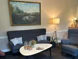 En hyggelig stue med en mørkegrå sofa, lænestol, et hvidt sofabord med snacks og blomster, en gulvlampe, en stueplante og et landskabsmaleri på væggen.