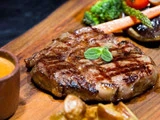 Grillet ribeye-bøf med grillmærker, toppet med friske krydderurter, serveret på et træbræt med ristede grøntsager og en dypsauce.