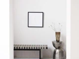 Minimalistisk indretning med en stribet bænk, et rundt sidebord i metal, en vase med tørrede blomster og et indrammet kunstværk på en hvid væg.