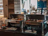 To buffetvarmere i rustfrit stål fyldt med forskellige varme retter er opstillet på en disk i en restaurant. Tallerkener og redskaber er placeret foran.