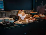 En morgenmadsbuffet med forskellige brød, croissanter, kager, en brødrister, tallerkener, smør og ost på en mørk disk mod en rustik trævæg.