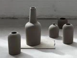 Fem matte, minimalistiske keramikvaser i neutrale toner er arrangeret på et betongulv, og en vase er placeret på en rektangulær stenplade.