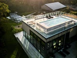 En moderne bygning med en pool på taget omgivet af glasrækværk, liggestole og drivhuse i nærheden, set i sollyset.