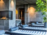 En moderne spa-lounge med polstrede lænestole, en glaspejs, grå flisevægge, dæmpet belysning og en grøn plante.