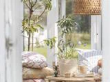 En hyggelig udestue med et lille bord, potteblomster, stablede puder, en kurvelampe og sollys, der strømmer ind gennem store vinduer.