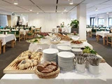 Et buffetbord med tallerkener, bestik, brød, frugt og salater i et rummeligt spiseområde med pænt opstillede borde og stole.