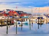 Farverige både ligger fortøjet i en marina, hvor det rolige vand spejler deres silhuetter. I baggrunden står røde bygninger under en delvis overskyet himmel ved solnedgang.