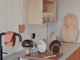 Minimalistisk køkkenbord med tørre tallerkener, glas og redskaber. Skærebrætter af træ, en brun vase og et håndklæde er også synlige ved siden af en sort vandhane og en træhylde.