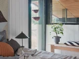 Et hyggeligt soveværelse med en seng, puder, en bordlampe, en potteplante på en træbænk og store vinduer, der lukker naturligt lys ind.