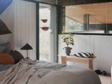 Et hyggeligt soveværelse med en uredt seng, en rund lampe, en potteplante på et træbord og store vinduer, der lukker naturligt lys ind.