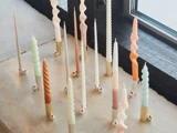 En gruppe pastelfarvede, høje, snoede stearinlys i guldlysestager står opstillet på en vindueskarm ved siden af et vindue med sort ramme.