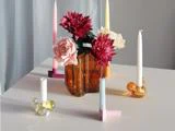 En brun glasvase med kunstige blomster og fire farverige geometriske lysestager med stearinlys på et hvidt bord.