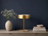 En guldfarvet bordlampe står på et beige skænk ved siden af en vase med hvide blomster, en stak bøger og en lille skål, med en mørkeblå væg i baggrunden.