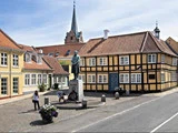 Et brostensbelagt torv med en statue af en kvinde i traditionel kjole, omgivet af farverige historiske bygninger og to mennesker i nærheden. Et kirkespir rejser sig i baggrunden.