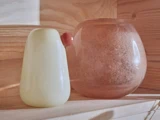 To glasgenstande på en træhylde: en glat, lysegul vase til venstre og en rund, gennemskinnelig pink vase med en lille knop til højre.