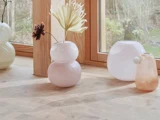 Tre pastelfarvede glasvaser, den ene med blomster, står i en vindueskarm af træ med udsigt til træer og blade udenfor.