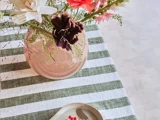 En rund lyserød vase med forskellige blomster står på en grøn- og hvidstribet dug ved siden af et lille fad med røde bær på en lys overflade.