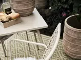 En hvid metalstol ved siden af et bord med brød og et stearinlys, placeret på et mønstret grønt udendørs tæppe med store potteplanter i nærheden.