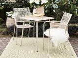 To hvide metalstole og et bord med mad og drikke står på et mønstret tæppe i en have med frodige planter og hvide blomster. Den ene stol har en hvid, blød hynde.