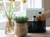 En træpotte med grønne krydderurter, en vase med orange og gule blomster og en sort kurv med krydderier og redskaber står på et træbord ved et vindue.