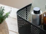 En sort plastkurv med huller indeholder flasker og en notesbog på en lys træoverflade ved siden af en grøn potteplante.