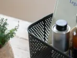 En sort perforeret kurv med en bog, en metaldunk og en brun flaske står på en træplade ved siden af en grøn potteplante.