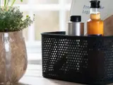 En sort perforeret kurv med flasker står på et bord ved siden af en potteplante, med et vindue og en sløret baggrund bag dem.