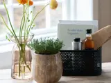 Et træbord med en vase med gule blomster, en potteplante og en sort kurv med flasker, en bog og redskaber ved et lyst vindue.