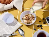 En hånd hælder mælk i en skål med granola på et bord med gule fliser, med en ske, en avis, blåbær, te og en croissant i nærheden.