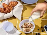 En hånd hælder mælk i en skål med granola på et gult kakkelbord med croissanter, blåbær, en avis og tomme skåle i nærheden.