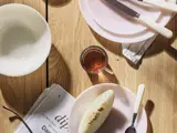 Et træbord dækket med hvide tallerkener, kniv og gaffel, en halveret melon, et glas te og en avis, der ligger delvist synlig i hjørnet.