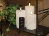 På en mørk hylde ved siden af en potteplante står tre hvide flammeløse stearinlys med lysende falske flammer og en sort fjernbetjening.