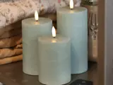 Tre lyseblå bloklys med glødende flammer er arrangeret sammen på en hylde med foldet stof og en dekorativ vase i baggrunden.