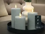 Tre grå bloklys med realistiske flammer er placeret på en sort bakke ved siden af en hvid vase. En fjernbetjening med knapper er synlig i forgrunden.
