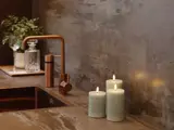 Tre tændte bloklys står på et moderne badeværelsesbord ved siden af en kobbervandhane med en sæbedispenser i glas og en lille vase med grønne planter i baggrunden.
