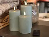 Tre blå bloklys med glødende flammer står på et bord ved siden af foldet sengetøj, en keramikvase og en sort fjernbetjening.