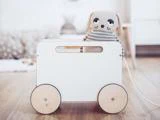 En blød, stribet tøjhund sidder i en hvid legetøjsvogn af træ med store runde hjul på et trægulv i et lyst rum.