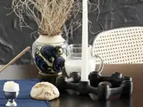 Et bord med en lysestage, et hvidt lys, en bolle, et blødkogt æg, en blå serviet og en kande mælk ved siden af en vase med tørrede planter.