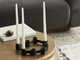 En moderne sort lysestage med fire høje hvide lys står på et rundt guldbord ved siden af en sofa og en bog med en figen på toppen.