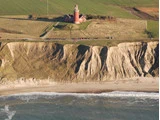 Et rødt fyrtårn står nær kanten af en græsklædt klippe med udsigt over havet, med sandstrand og bølger nedenfor.