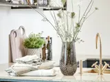 Et køkkenbord med en glasvase med høje hvide blomster, foldede håndklæder, krydderurter i potter, skærebrætter og flasker ved siden af en guldvandhane.